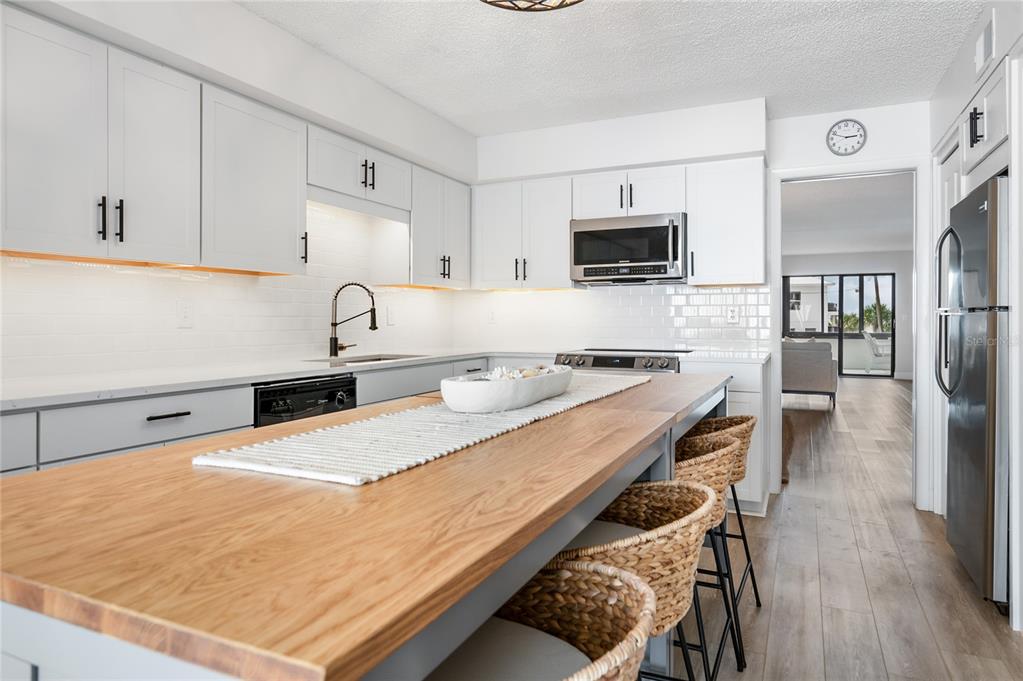 A kitchen features white cabinets, a wooden island with woven stools, and stainless steel appliances — 1007 GULF DRIVE N UNIT 106, BRADENTON BEACH, FL