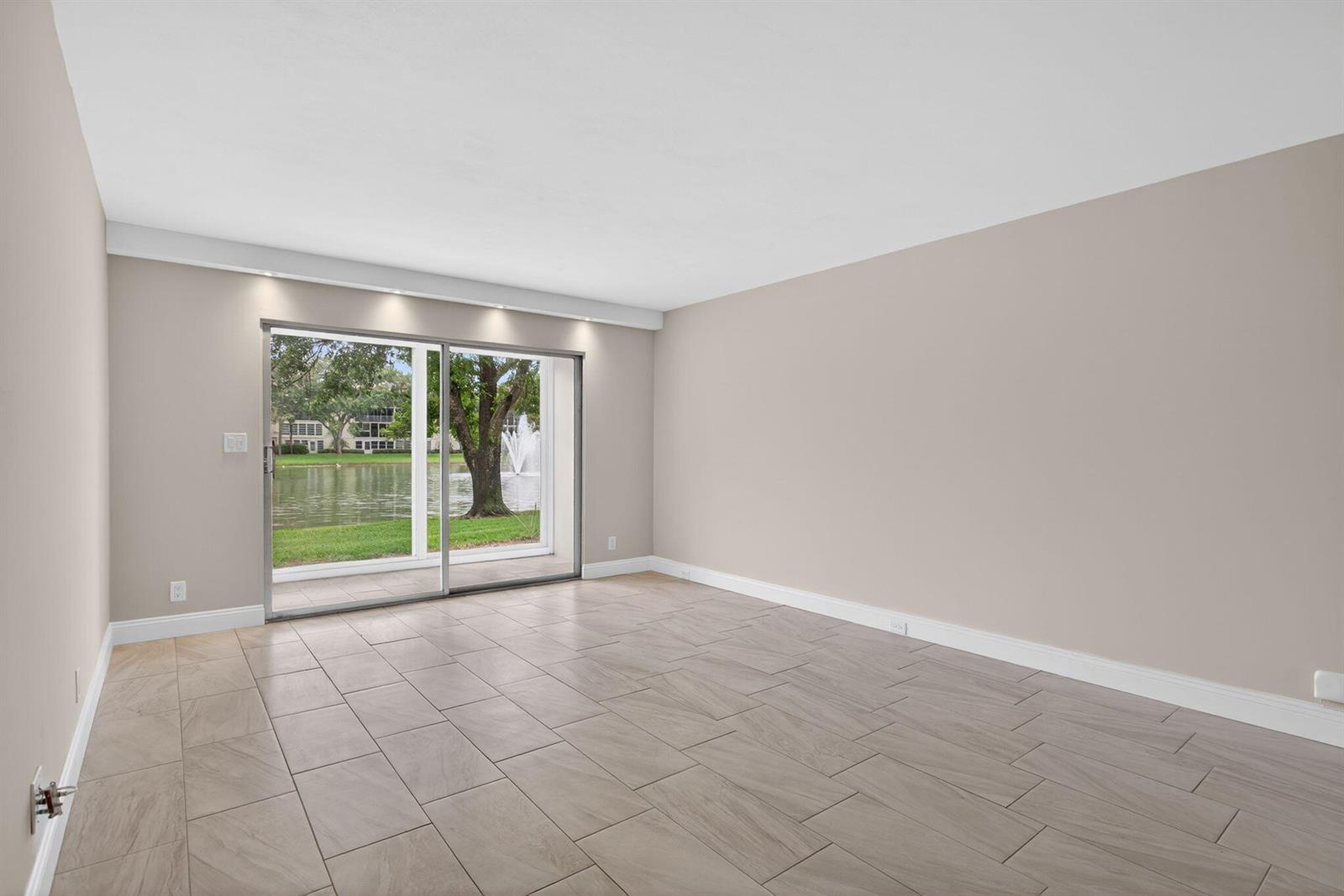 An empty room features light-colored tile flooring, beige walls, and a sliding glass door view — 2202 Lucaya Bend # B1, Coconut Creek FL 33066, COCONUT CREEK, FL
