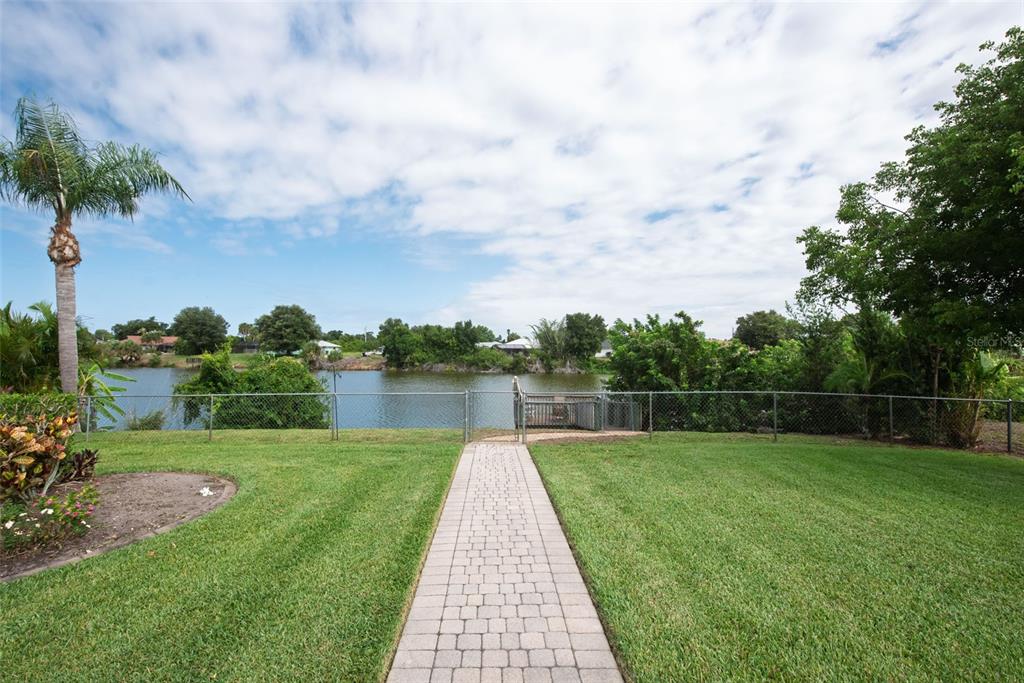 A paved walkway leads through a grassy backyard to a chain-link fence overlooking a lake — 7411 SNOW DRIVE, ENGLEWOOD, FL