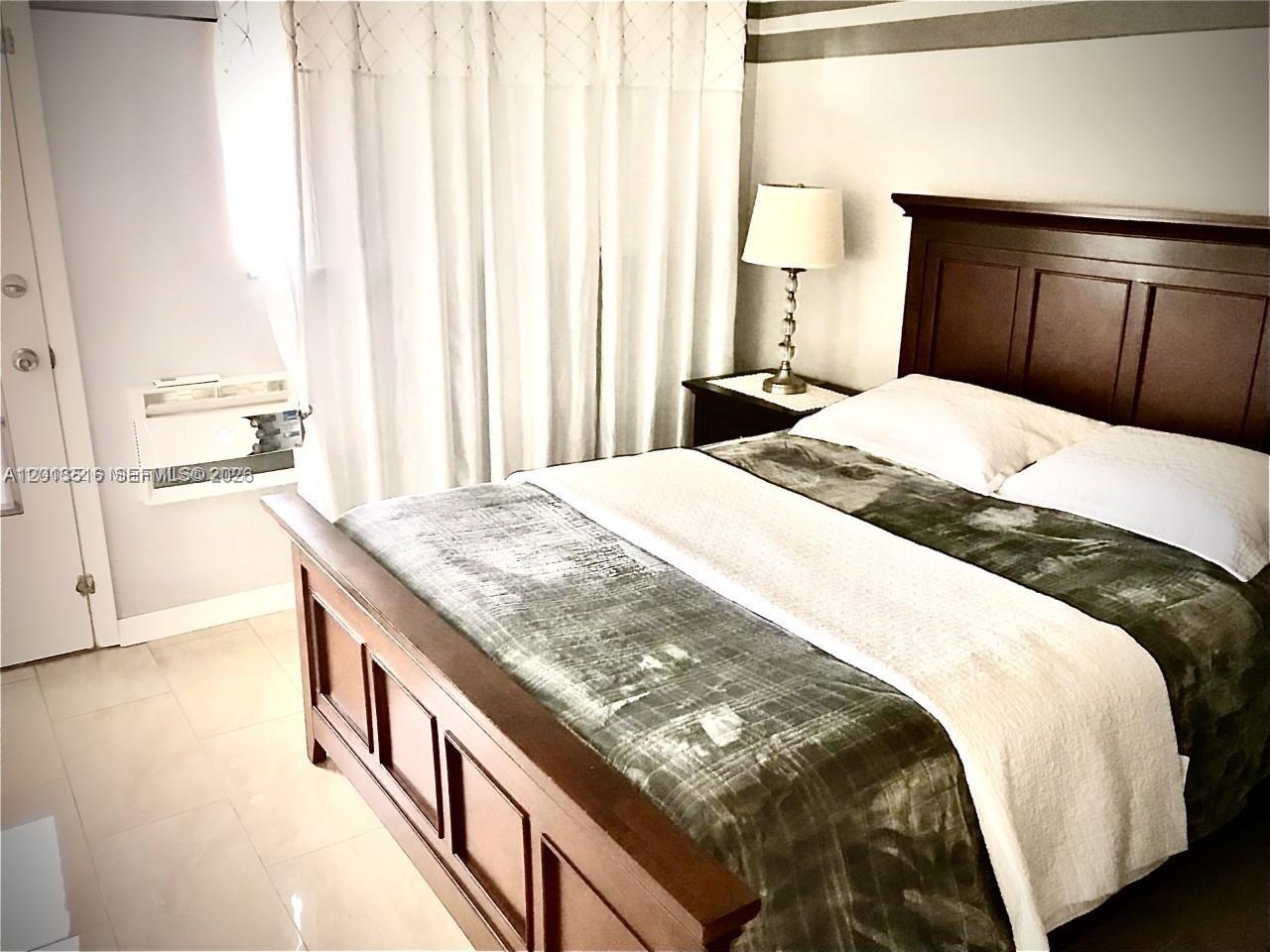 A bedroom features a dark wood bed frame, a nightstand with a lamp, and white curtains — 900 SW 11th Ave # 17C, Hallandale Beach FL 33009, HALLANDALE BEACH, FL