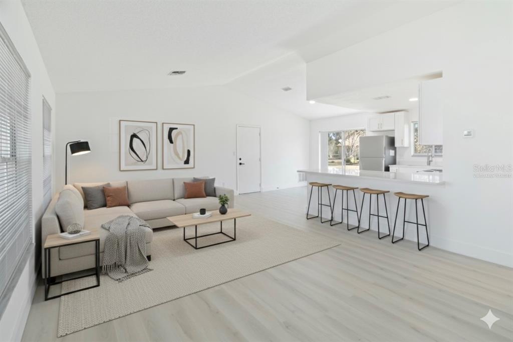 This virtually staged living room features light wood flooring, a sectional sofa, and kitchen seating — 11407 SPLITWOOD LANE, ORLANDO, FL