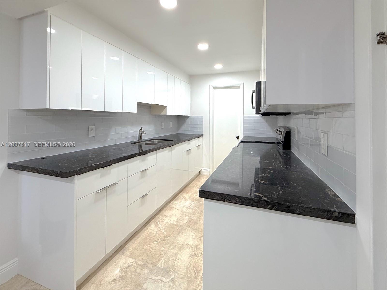 A galley kitchen features white high-gloss cabinets, dark stone countertops, and light tile flooring — 1075 92nd St # 201, Bay Harbor Islands FL 33154, BAY HARBOR ISLANDS, FL