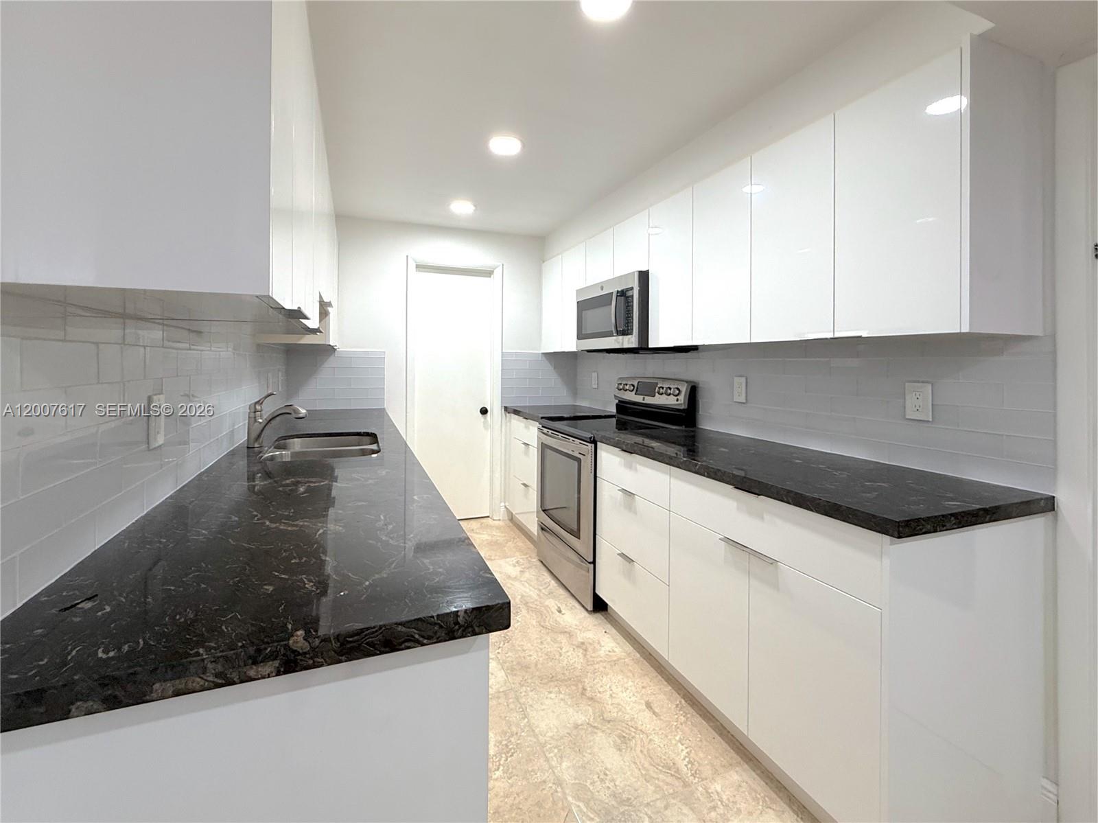 A galley kitchen features white cabinets, black countertops, stainless steel appliances, and light tile flooring — 1075 92nd St # 201, Bay Harbor Islands FL 33154, BAY HARBOR ISLANDS, FL