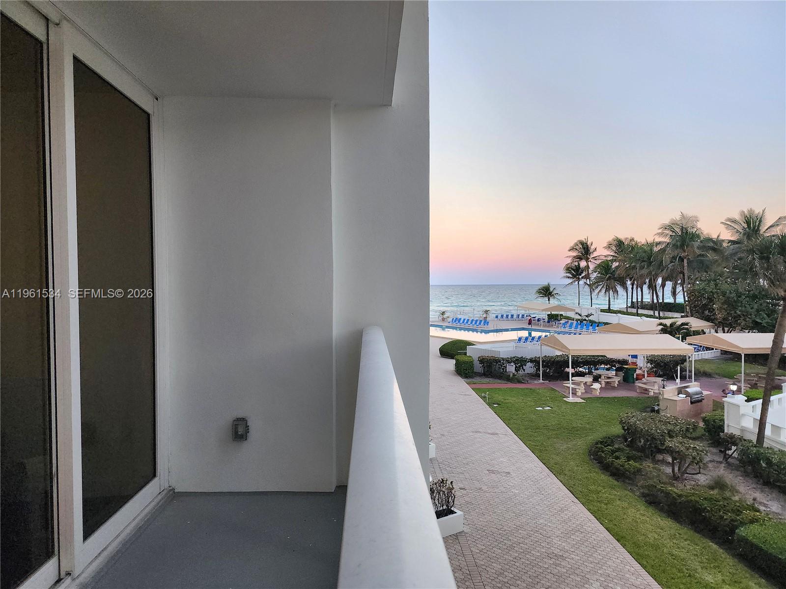 A balcony overlooks a resort pool area, palm trees, and the ocean at sunset — 3505 S Ocean Dr # 218, Hollywood FL 33019, HOLLYWOOD, FL