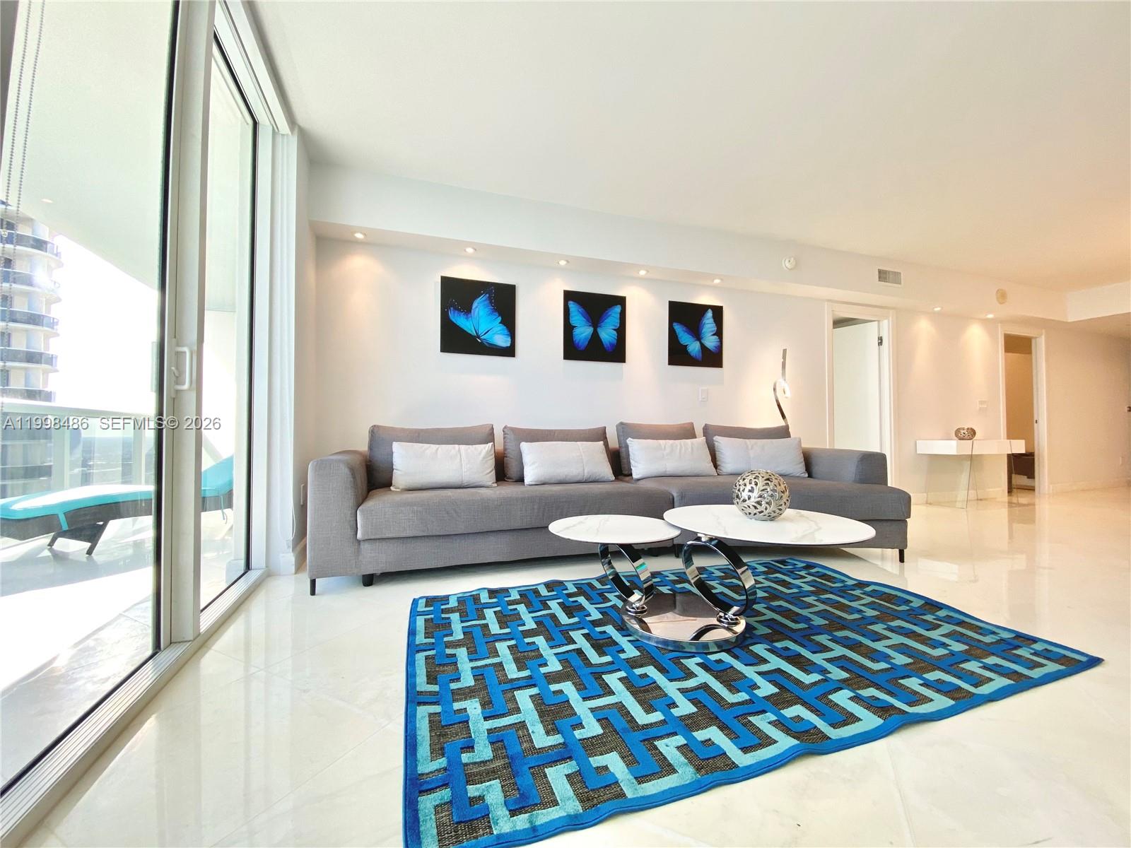 A living room features a grey sectional sofa, a patterned blue rug, and butterfly wall art — 1830 S Ocean Dr # 3203, Hallandale Beach FL 33009, HALLANDALE BEACH, FL
