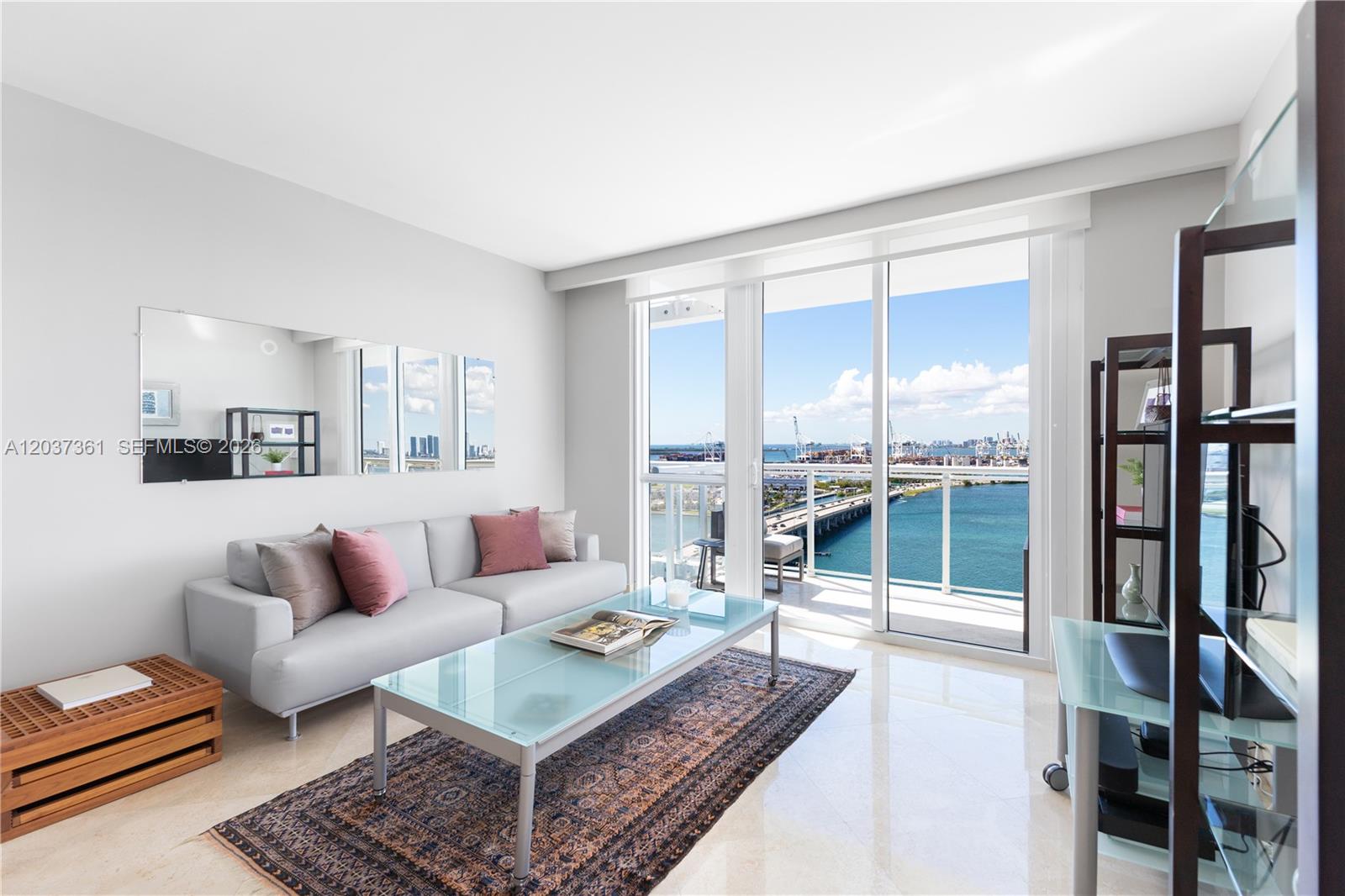 A living room features a gray sofa, glass coffee table, and a balcony overlooking water — 520 West Ave # 2004, Miami Beach FL 33139, MIAMI BEACH, FL