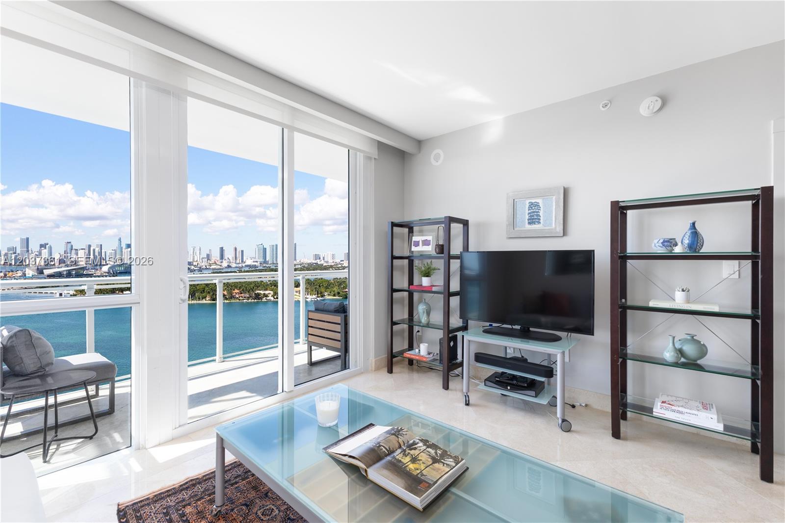 A living room features a glass coffee table, shelving units, and a balcony with water views — 520 West Ave # 2004, Miami Beach FL 33139, MIAMI BEACH, FL