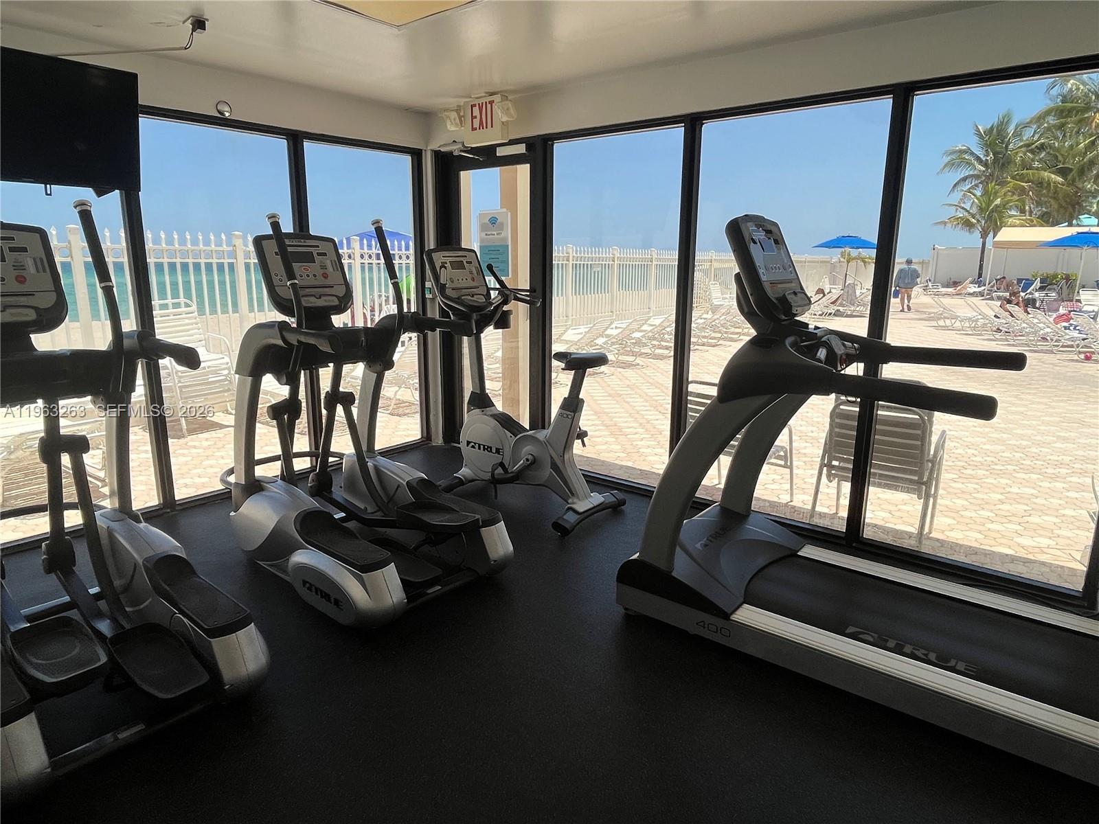 A gym features exercise equipment facing floor-to-ceiling windows overlooking a pool deck and beach — 3505 S Ocean Dr # 502, Hollywood FL 33019, HOLLYWOOD, FL