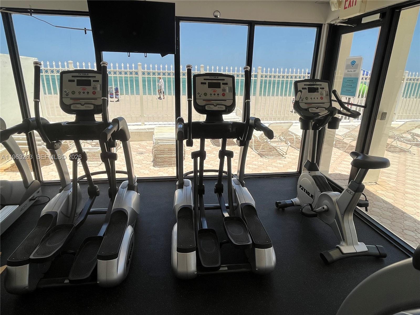 A gym room features three exercise machines facing a large window overlooking a sandy beach — 3505 S Ocean Dr # 502, Hollywood FL 33019, HOLLYWOOD, FL