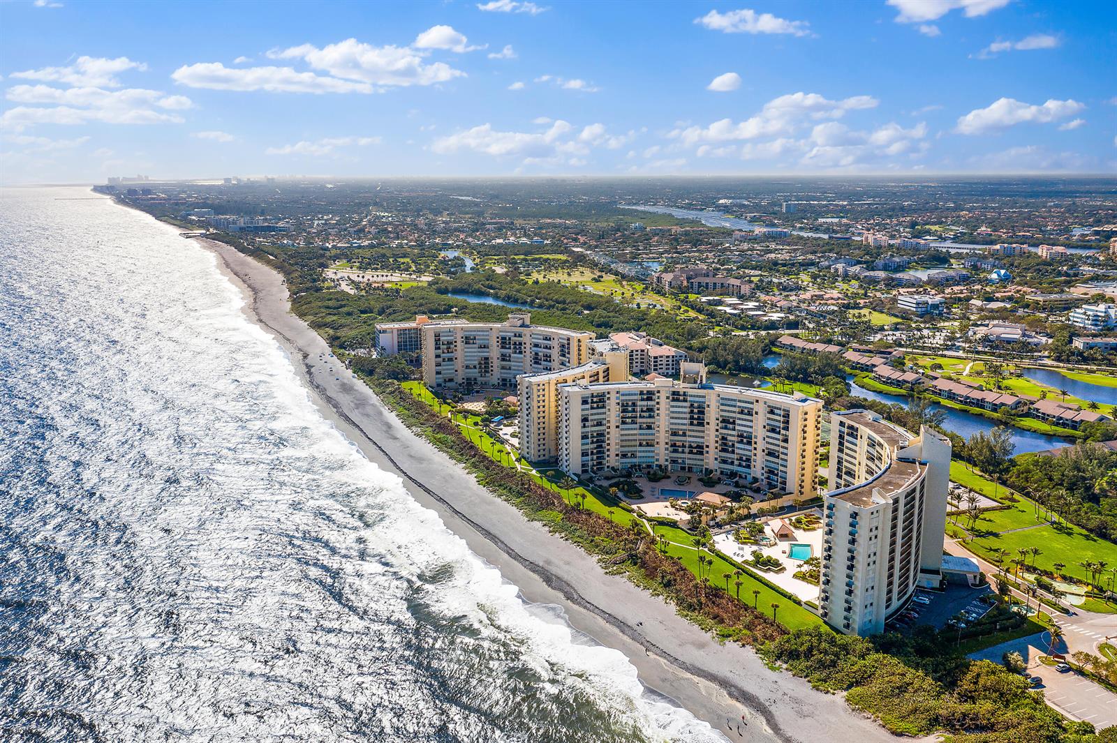 An aerial view shows curved beachfront condominium buildings along a coastline with surrounding green space — 300 Ocean Trail Way # 403, Jupiter FL 33477, JUPITER, FL
