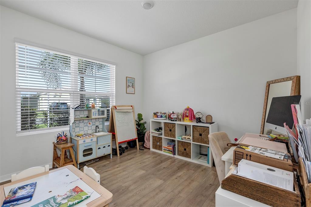 A bright playroom features wood-look flooring, a toy kitchen, a desk, and white shelving units — 3221 SILAS DRIVE, NEW SMYRNA BEACH, FL