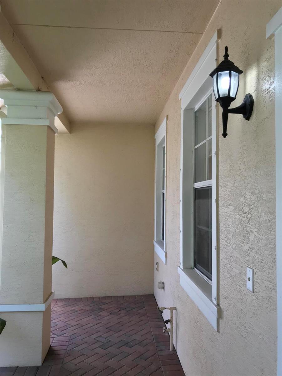 A covered porch features a brick floor, a beige stucco wall, and a wall-mounted light fixture — 141 E Thatch Palm Circle, Jupiter FL 33458, JUPITER, FL