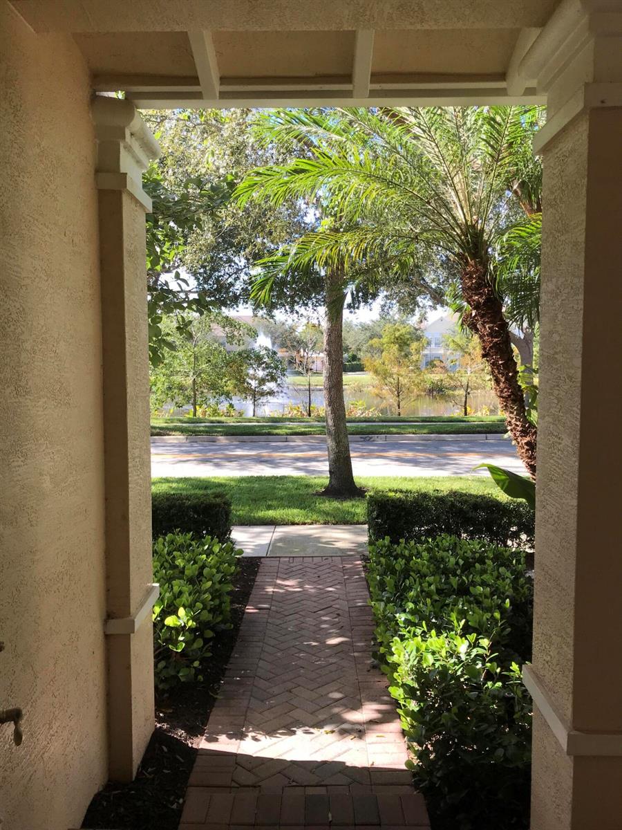 A view from a covered porch looking out toward a brick walkway, lawn, and pond — 141 E Thatch Palm Circle, Jupiter FL 33458, JUPITER, FL