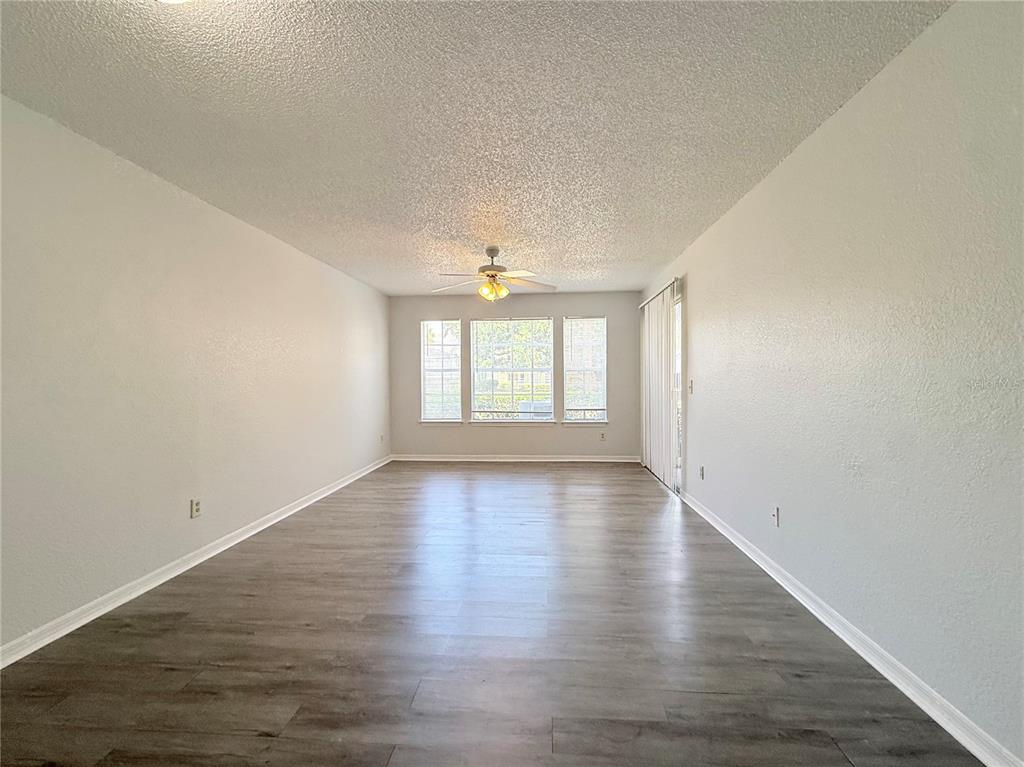 An empty living room features light-colored walls, wood-look flooring, a ceiling fan, and large windows — 1025 S HIAWASSEE ROAD UNIT 2216, ORLANDO, FL