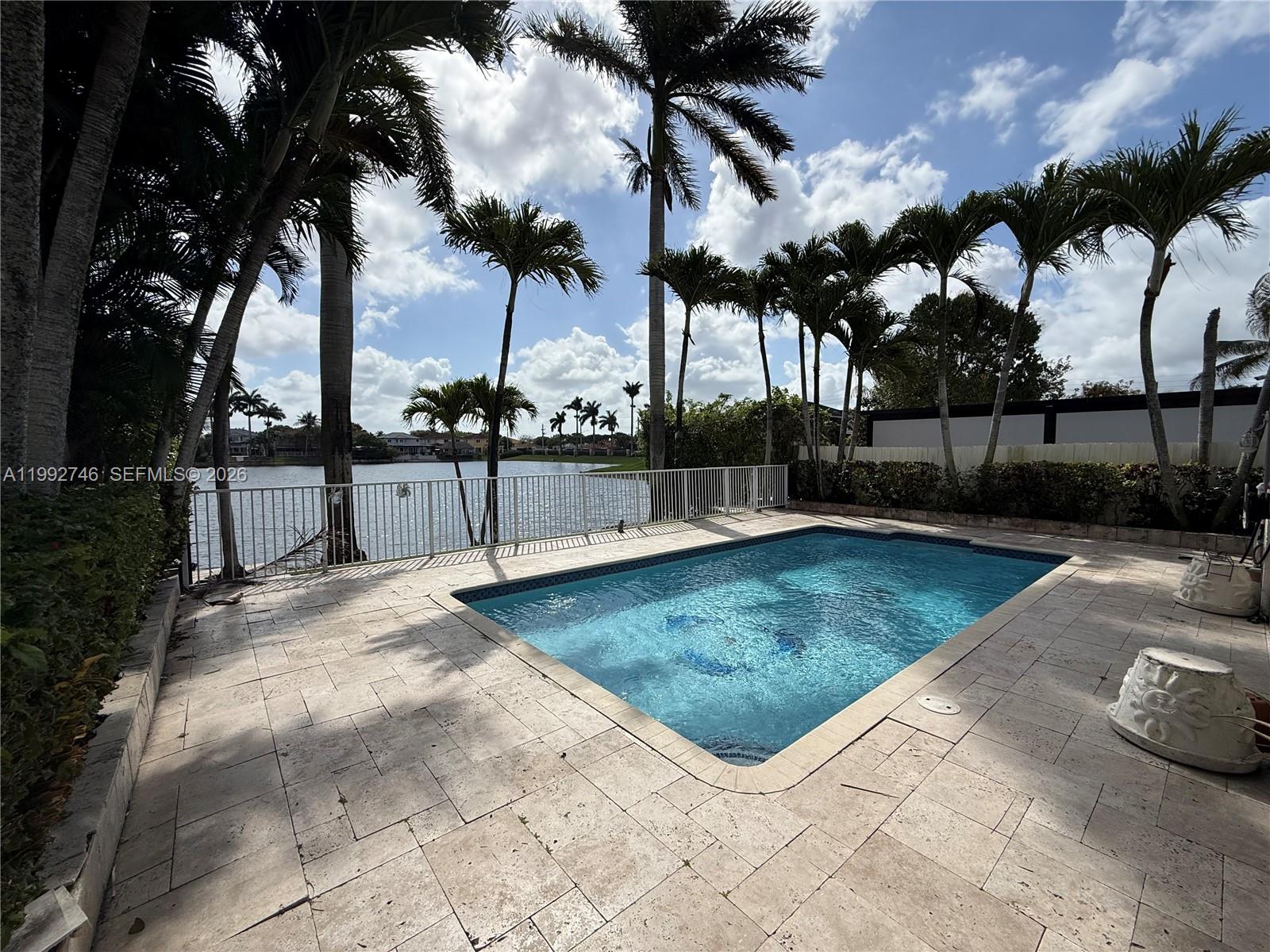 A rectangular swimming pool sits on a stone patio overlooking a lake with palm trees — 5513 SW 164th Ct, Miami FL 33185, MIAMI, FL