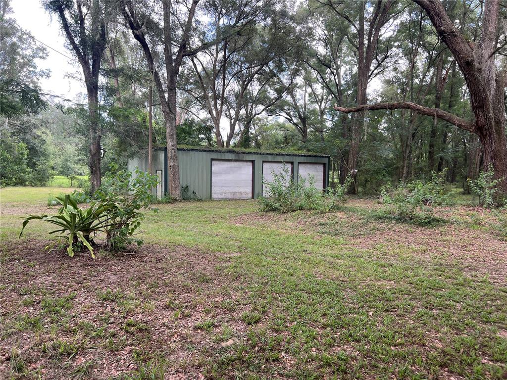 A detached metal garage with three roll-up doors sits in a wooded, grassy yard — 19107 SW 179 PLACE, ARCHER, FL