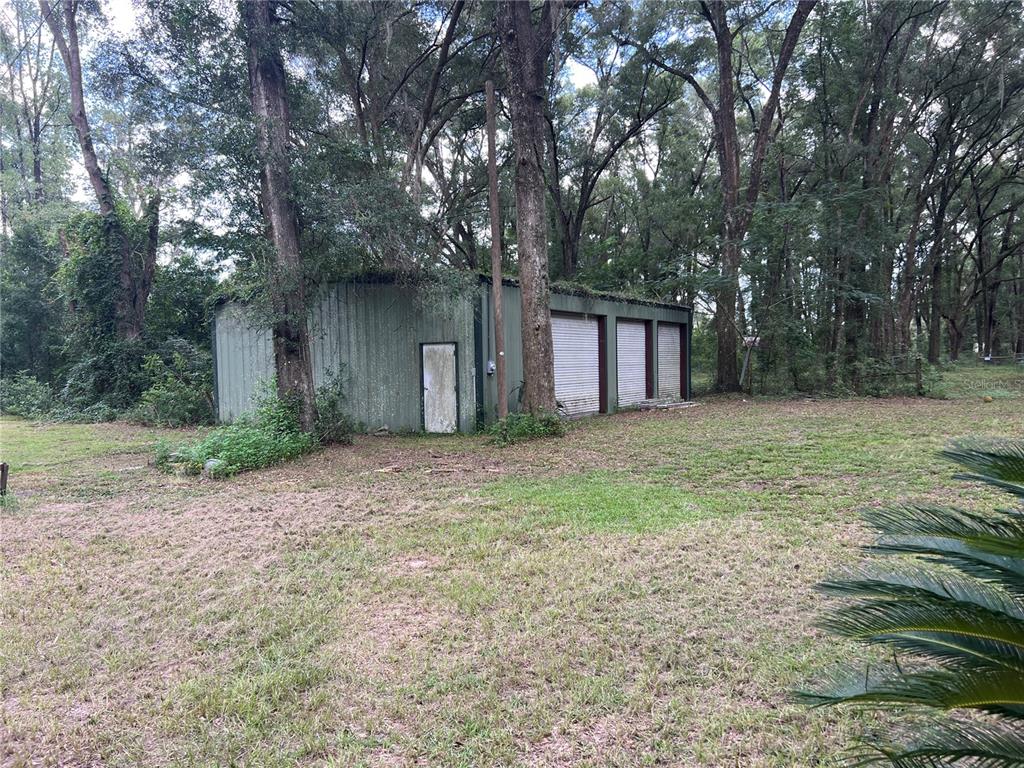 A green metal workshop with three roll-up doors stands in a wooded yard — 19107 SW 179 PLACE, ARCHER, FL