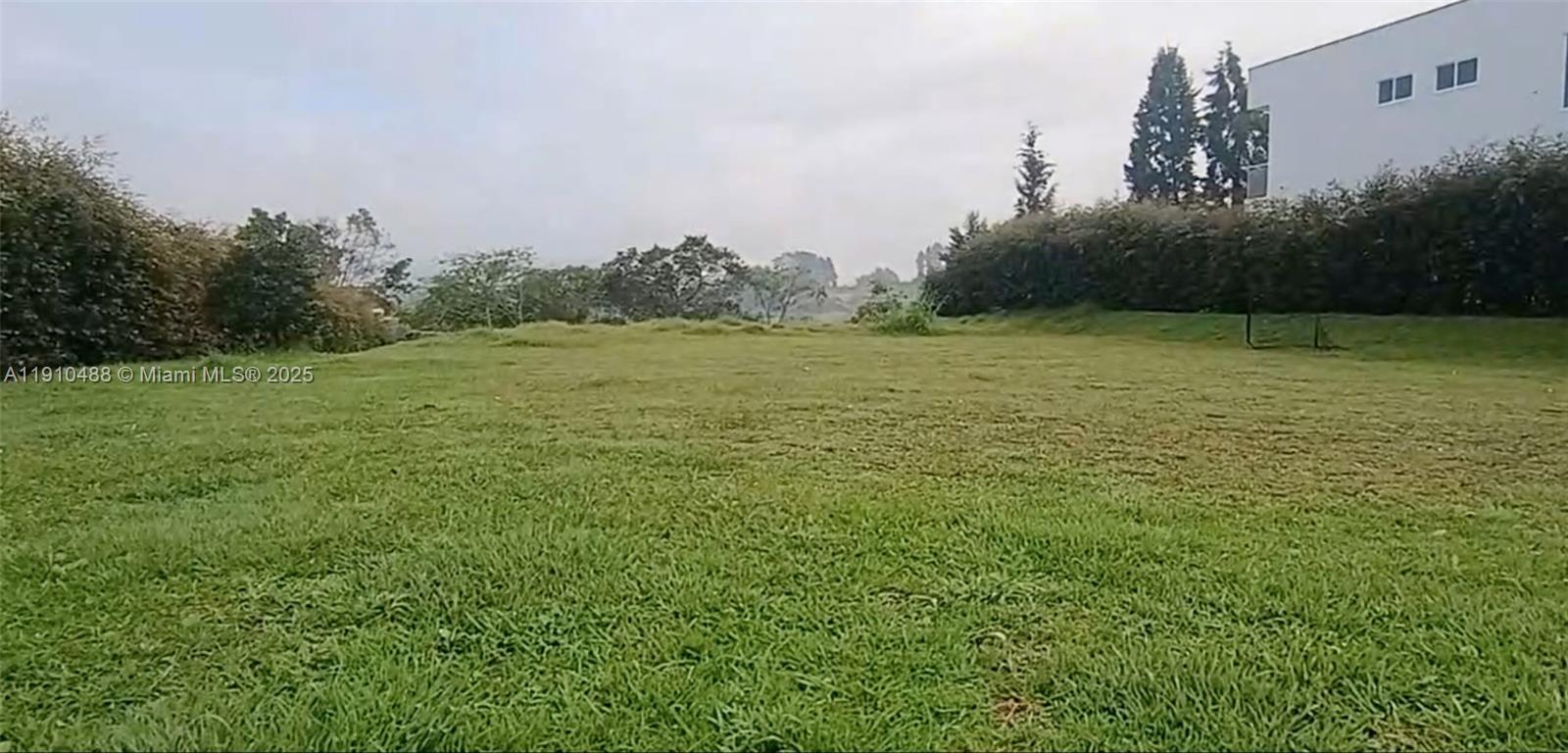 A grassy, open lot is bordered by trees and a modern white building — Vereda Mampuesto Sector Garrido #2, C Colombia, C