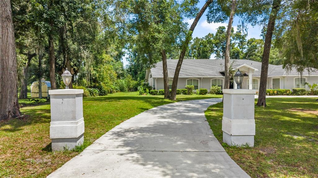 A concrete driveway leads to a single-story house framed by trees and two stone pillars — 11108 NW 70TH CIRCLE, CHIEFLAND, FL