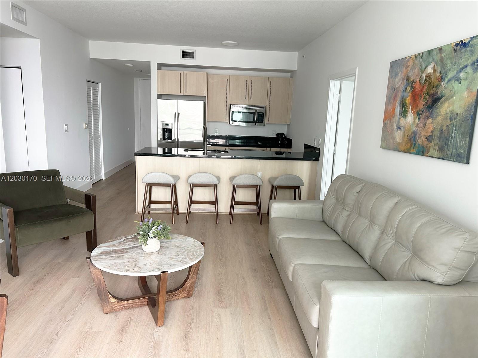 This living room features light wood flooring, a grey sofa, and an open kitchen area — 79 SW 12th St, Miami FL 33130, MIAMI, FL
