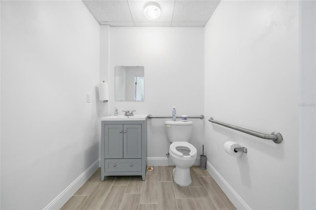 A small, white-walled bathroom features a grey vanity, a toilet with grab bars, and wood-look flooring — 30924 SUNEAGLE DRIVE UNIT Suite 201-203, MOUNT DORA, FL