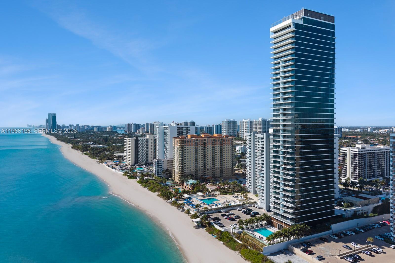 An aerial view shows a tall glass skyscraper on a sandy beach with surrounding buildings — 2000 S Ocean Dr # 25B, Hallandale Beach FL 33009, HALLANDALE BEACH, FL