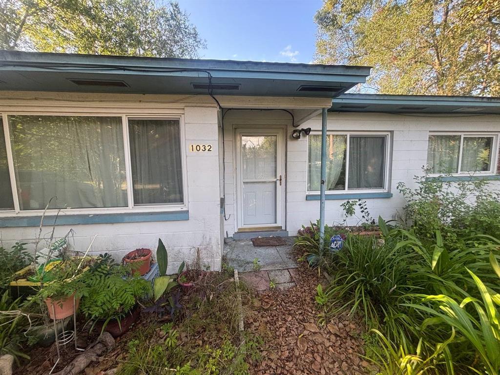 The exterior of a single-story white house features a front door, windows, and overgrown landscaping — 1032 NE 20TH AVENUE, GAINESVILLE, FL