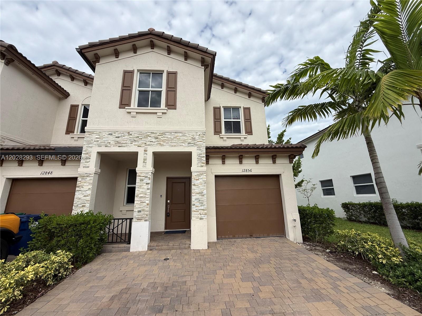 A two-story townhouse features a stone-accented facade, brown garage doors, and a paved driveway — 12856 NW 22nd Pl # 12856, Miami FL 33167, MIAMI, FL