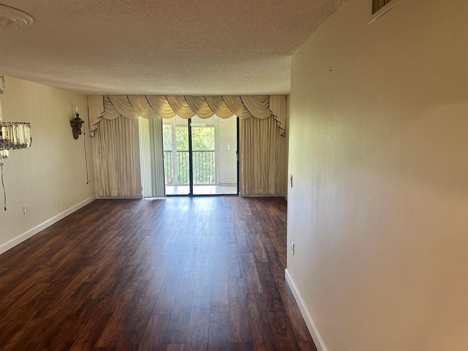 An empty living room features dark wood-look flooring, a sliding glass door, and decorative curtains — 3521 Environ Boulevard # B503, Lauderhill FL 33319, LAUDERHILL, FL