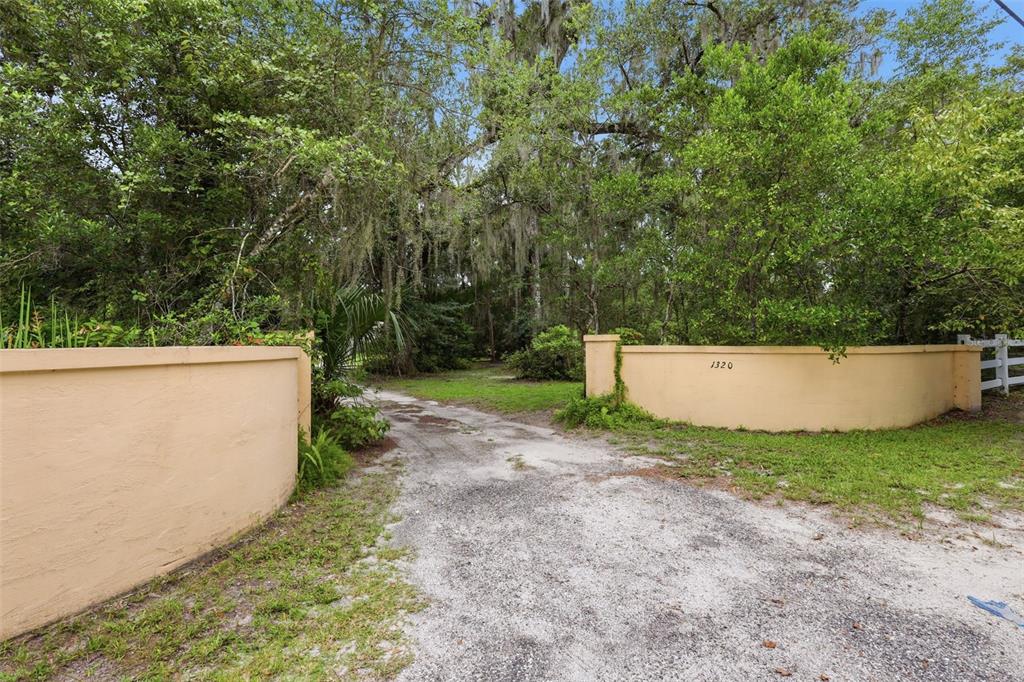 A gravel driveway leads through a gated entrance with tan stucco walls into a wooded area — 1320 W TAYLOR ROAD, DELAND, FL