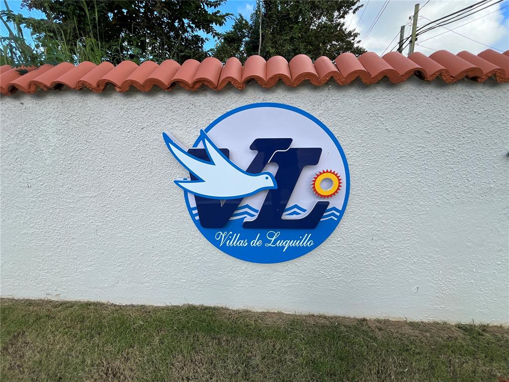 A white stucco wall features a circular logo for Villas de Luquillo with a bird — Urb. Villas de Luquillo C 15, LUQUILLO, PR