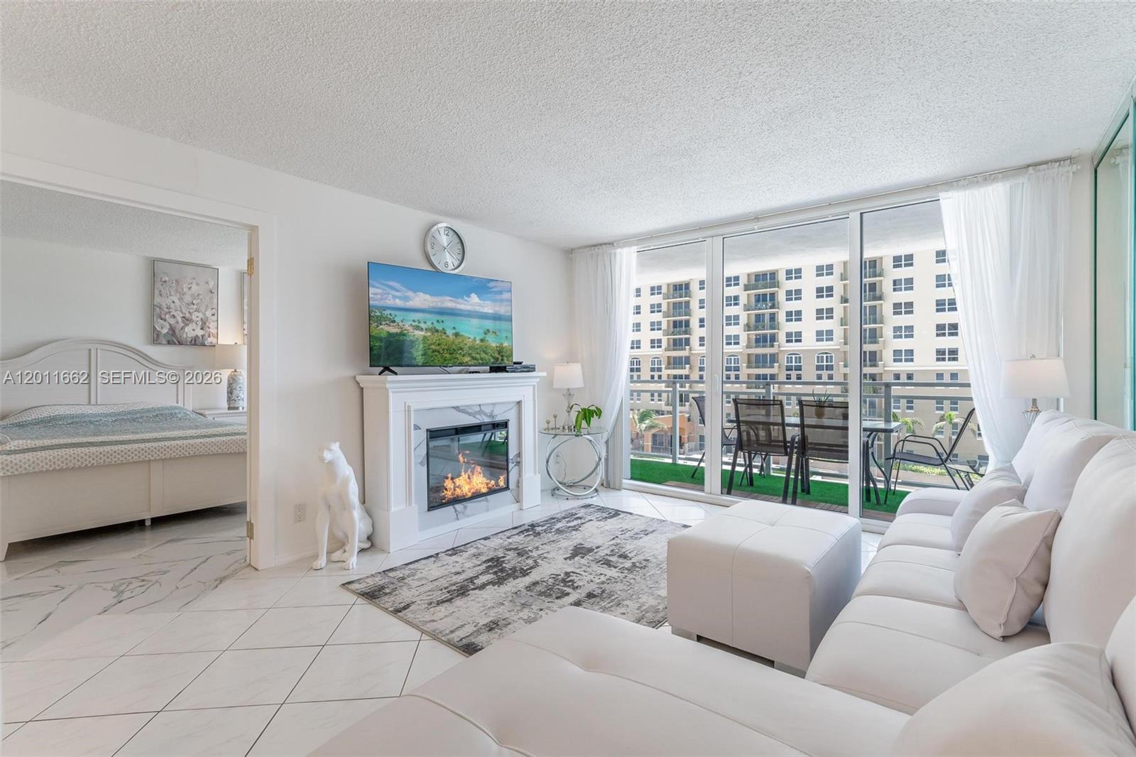A living room features a fireplace, a television, white furniture, and a balcony with seating — HALLANDALE BEACH, FL