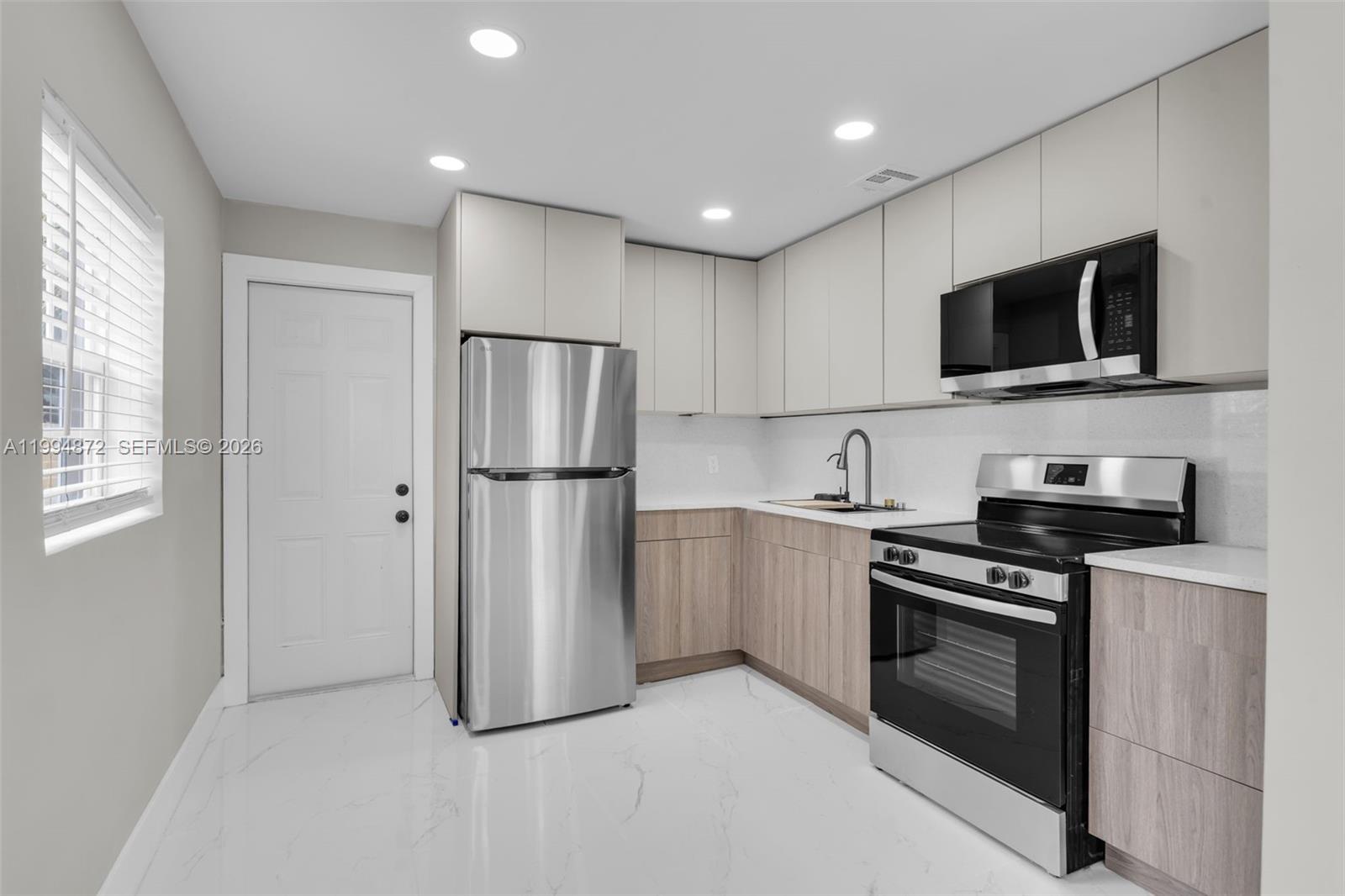 A modern kitchen features light wood cabinets, stainless steel appliances, and white marble-look flooring — 1660 W 26th St, Riviera Beach FL 33404, RIVIERA BEACH, FL