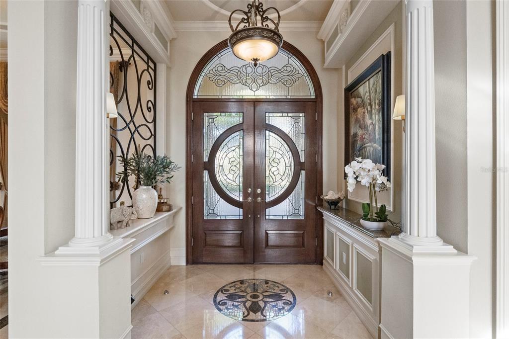 An elegant foyer features double wood doors, marble flooring, decorative columns, and a central chandelier — 7202 TEAL CREEK GLEN, LAKEWOOD RANCH, FL