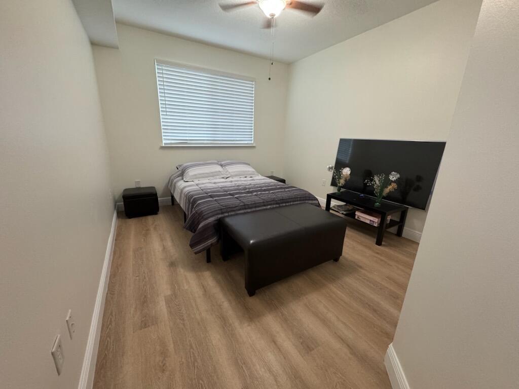 A bedroom with light wood-look flooring, a bed, a television on a stand, and blinds — 9873 Baywinds Boulevard # 5203, West Palm Beach FL 33411, WEST PALM BEACH, FL