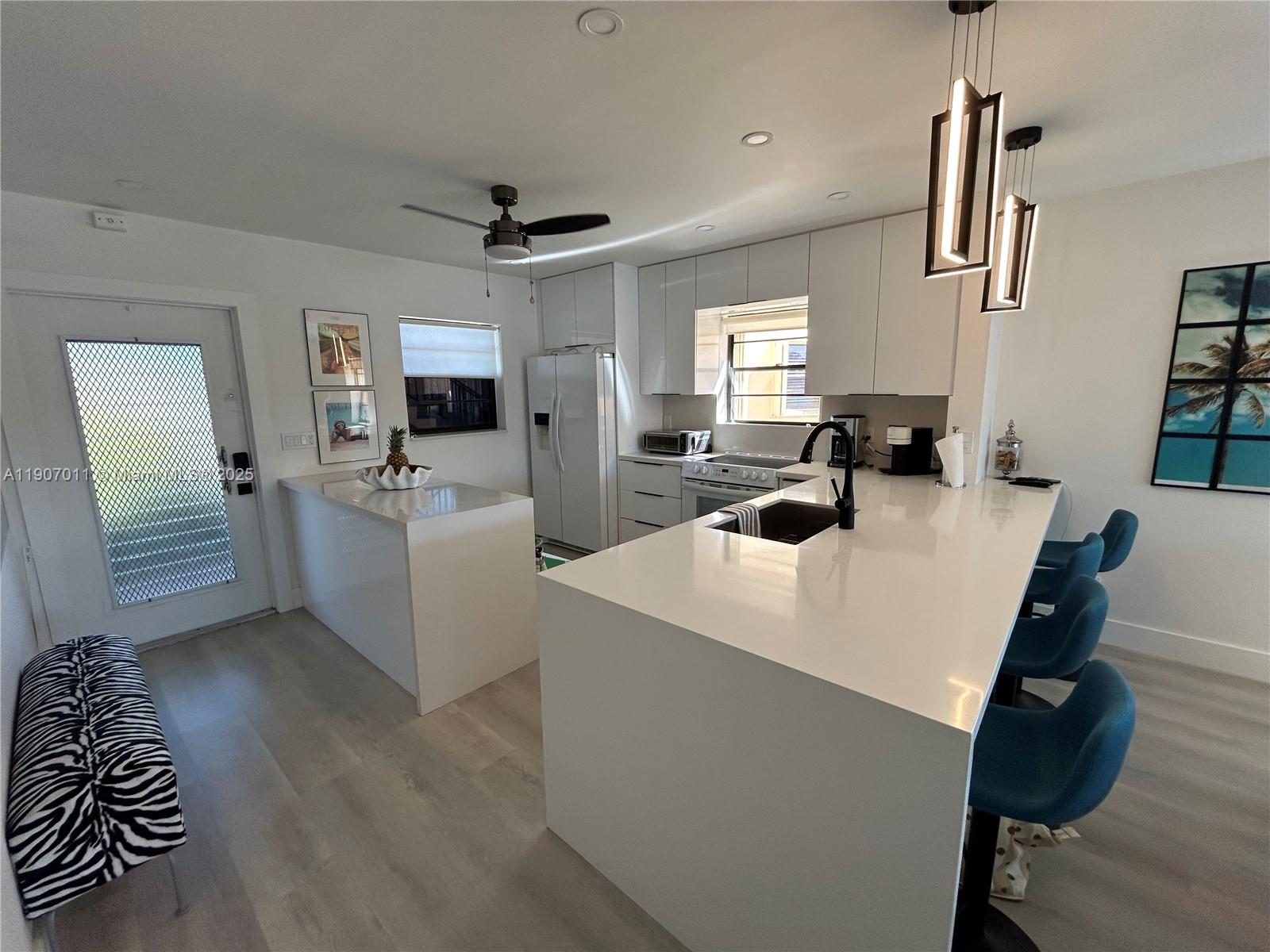A modern kitchen features white cabinetry, a large quartz island, and blue bar stools — 200 Berkley Rd # 113, Hollywood FL 33024, HOLLYWOOD, FL