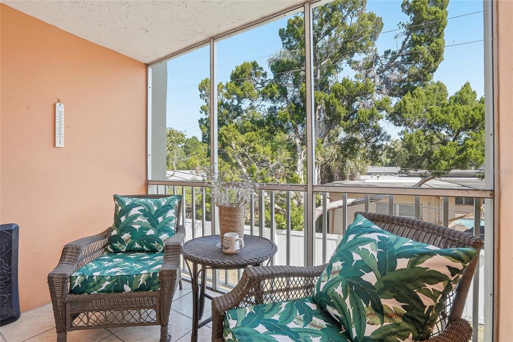 A wicker chair with palm-print cushions sits next to a small table on a balcony — 952 VIRGINIA STREET UNIT 202, DUNEDIN, FL