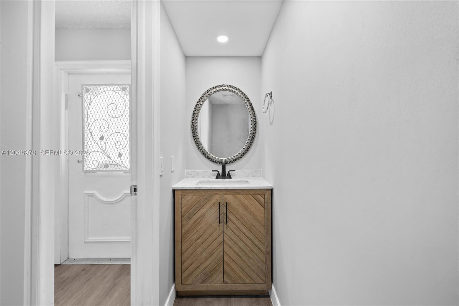 A small bathroom features a wood vanity, a round mirror, and a white door — 2408 NE 10th St # 122, Hallandale Beach FL 33009, HALLANDALE BEACH, FL