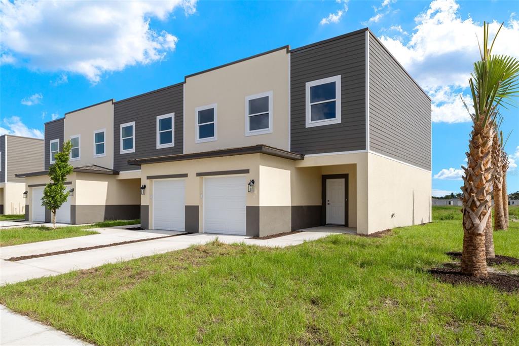 A two-story townhouse exterior with a tan and gray facade, two garage doors, and landscaping — 14985 SUNSET EVE LOOP, FORT MYERS, FL