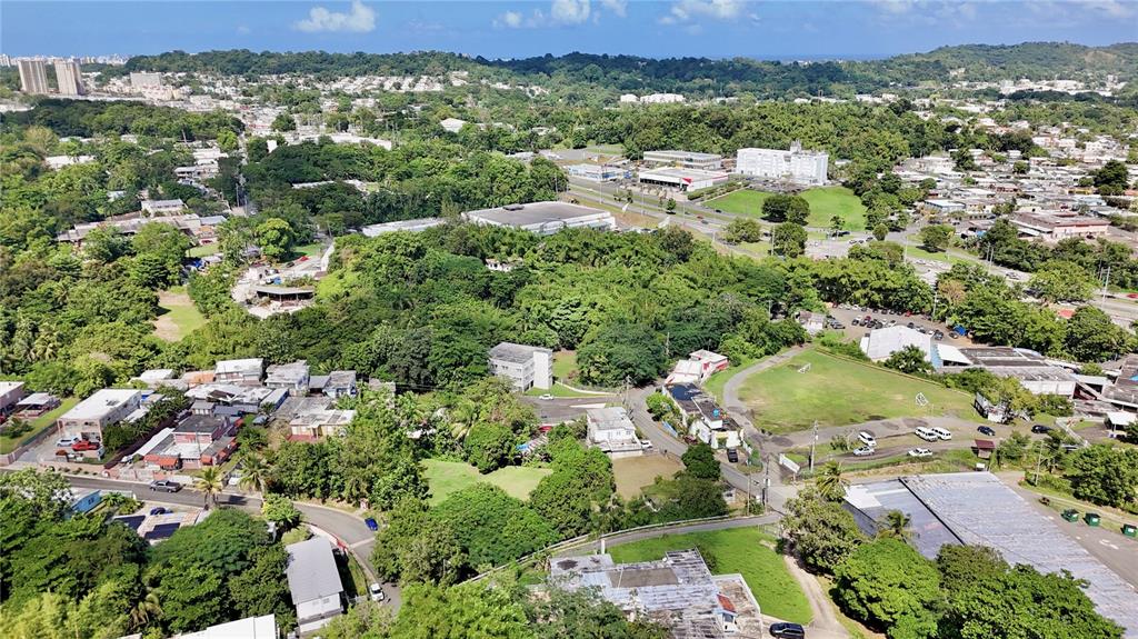 An aerial view shows a residential neighborhood with scattered buildings, lush green trees, and roads — PR-876 KM 0.7 BO LAS CUEVAS, TRUJILLO ALTO, PR