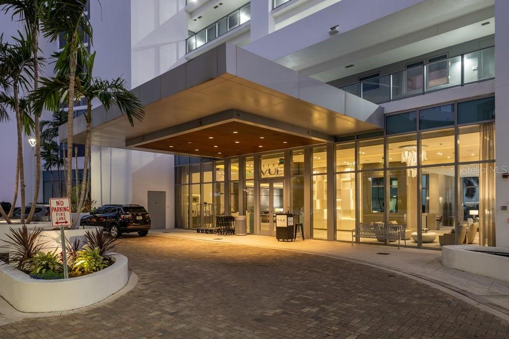 A modern apartment building entrance features a covered driveway, glass facade, and palm trees — 1155 N GULFSTREAM AVENUE UNIT 802, SARASOTA, FL