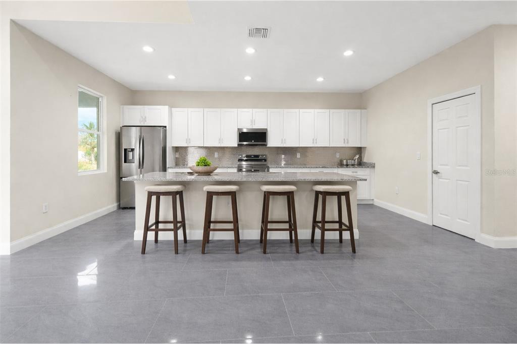 A modern kitchen features white cabinetry, a granite-topped island with four stools, and gray flooring — 1043 ALBERT AVE, LEHIGH ACRES, FL