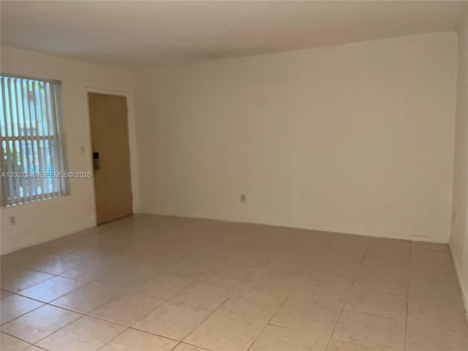 An empty room features light-colored tile flooring, white walls, and a window with vertical blinds — 255 Sunrise Dr # 103, Key Biscayne FL 33149, KEY BISCAYNE, FL