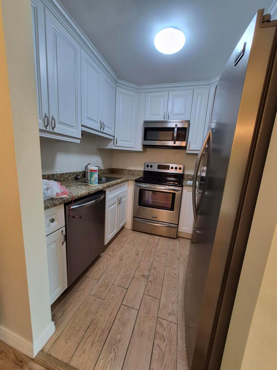 A galley kitchen features white cabinets, granite countertops, stainless steel appliances, and wood-look tile flooring — 27 Easthampton B, West Palm Beach FL 33417, WEST PALM BEACH, FL