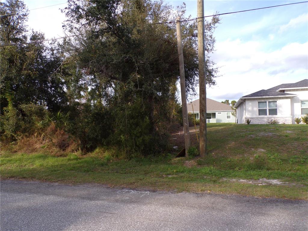 A vacant, tree-covered lot sits next to a suburban house with a paved road nearby — MEROLLA ROAD, NORTH PORT, FL