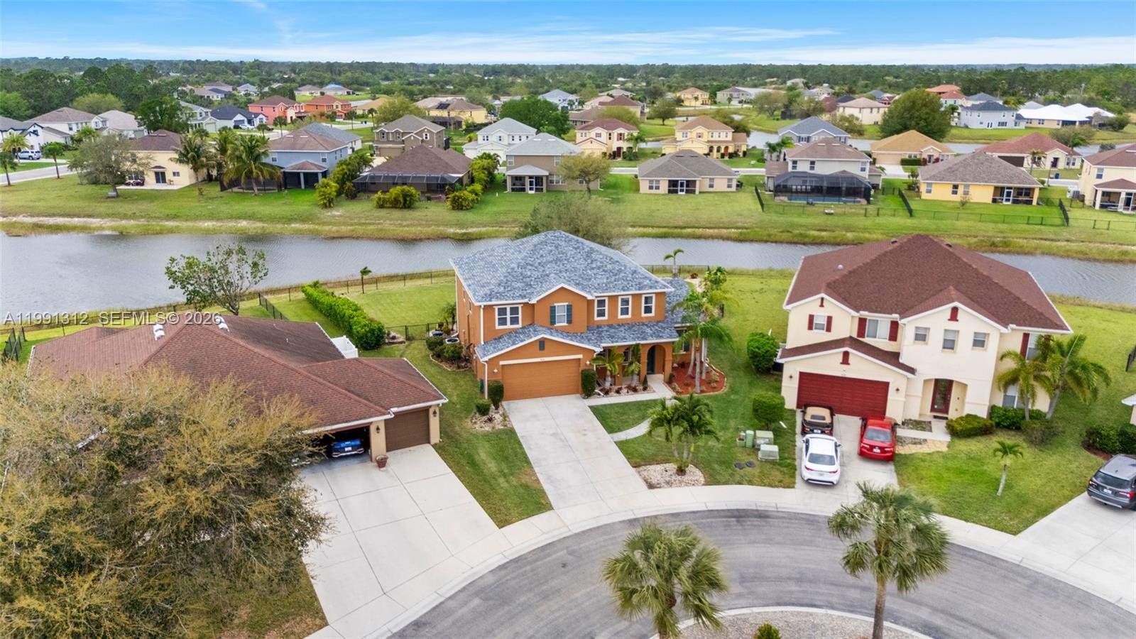 An aerial view shows three suburban houses with driveways and lawns bordering a small canal — 8130 blue daze ct, Lehigh Acres FL 33972, LEHIGH ACRES, FL