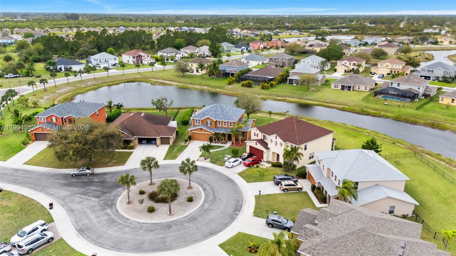 An aerial view shows a suburban neighborhood with houses surrounding a circular cul-de-sac and pond — 8130 blue daze ct, Lehigh Acres FL 33972, LEHIGH ACRES, FL