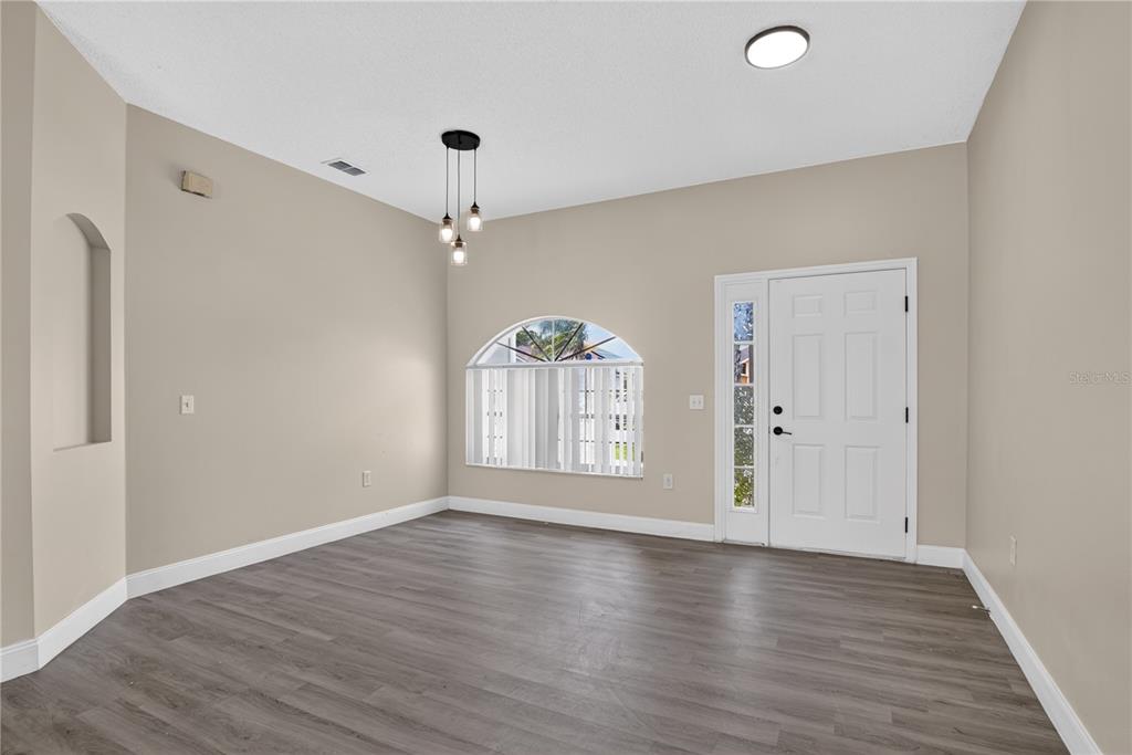 An empty room features gray wood-look flooring, beige walls, a front door, and a pendant light — 121 AURELIA COURT, KISSIMMEE, FL