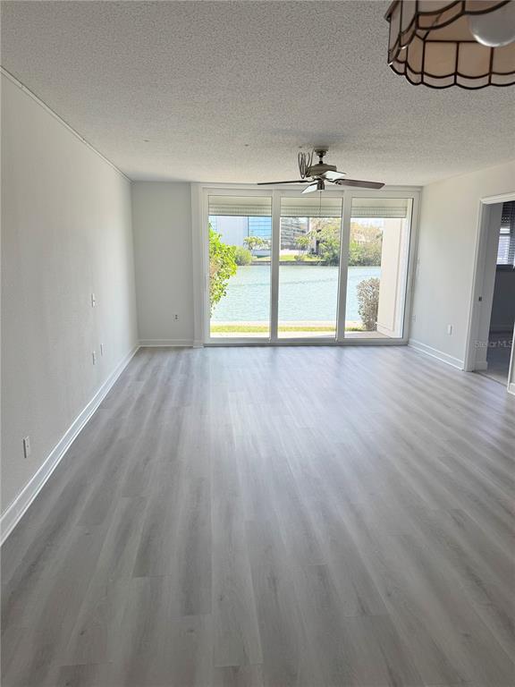 An empty living room with light wood-look flooring, a ceiling fan, and large windows overlooking water — 7425 BAY ISLAND DRIVE S UNIT 103, SOUTH PASADENA, FL