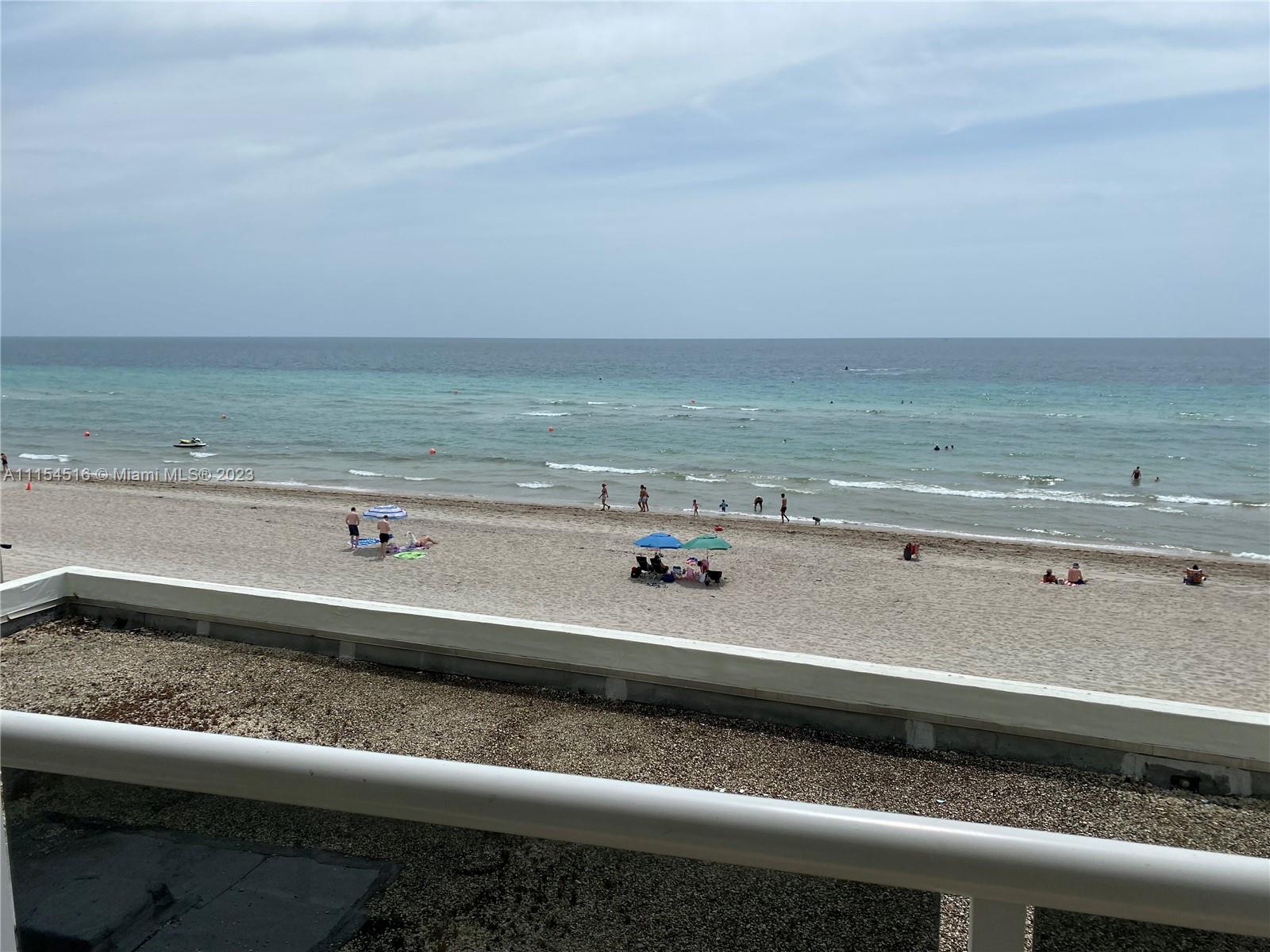 A balcony overlooks a sandy beach with people, umbrellas, and the ocean under a cloudy sky — 3725 S Ocean Dr # 301, Hollywood FL 33019, HOLLYWOOD, FL